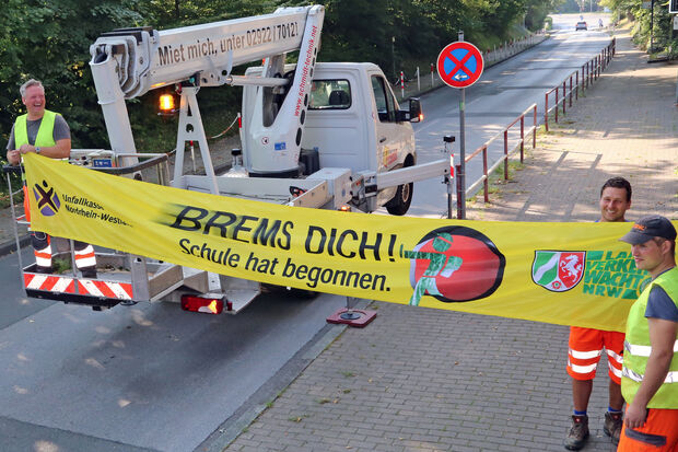 Mitarbeiter des kommunalen Bauhofes mit einem Hubwagen bei der Montage des leuchtend gelben Banners "BREMS DICH! Schule hat begonnen." der Landesverkehrswacht und der Unfallkasse Nordrhein-Westfalen an der Kirchstraße in Höhe der Engelhard-Grundschule: Frank Sattler, Jan Werrmann und Max Oeleker (von links). FOTO: ANDREAS DUNKER